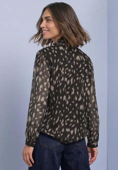 Femme de dos portant un chemisier noir à motifs beiges et un jean foncé, regard par-dessus l'épaule.