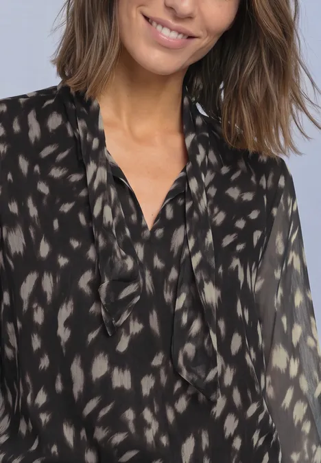 Une femme souriante porte une blouse noire transparente à motifs et col à volants.