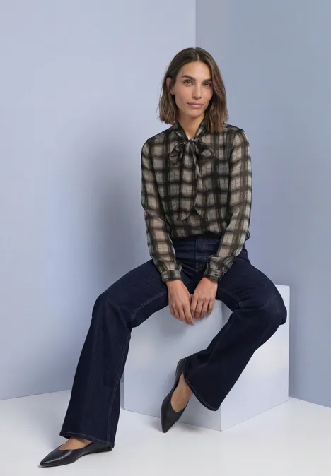 Chiffonblouse met strikdetail sanded amber Vrouw in geruit bloes met strik, donkere flared jeans en zwarte pumps, zittend op een grijs blok.