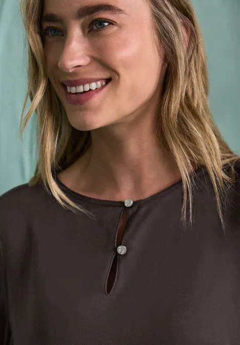 Chemisier à manches longues et à basque black coffee Gros plan d’une femme blonde souriante portant un haut marron orné de deux boutons au col.