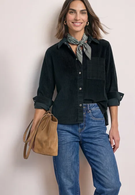 Frau mit schwarzem Hemd, blauen Jeans und brauner Umhängetasche, trägt ein gemustertes Halstuch und lächelt.