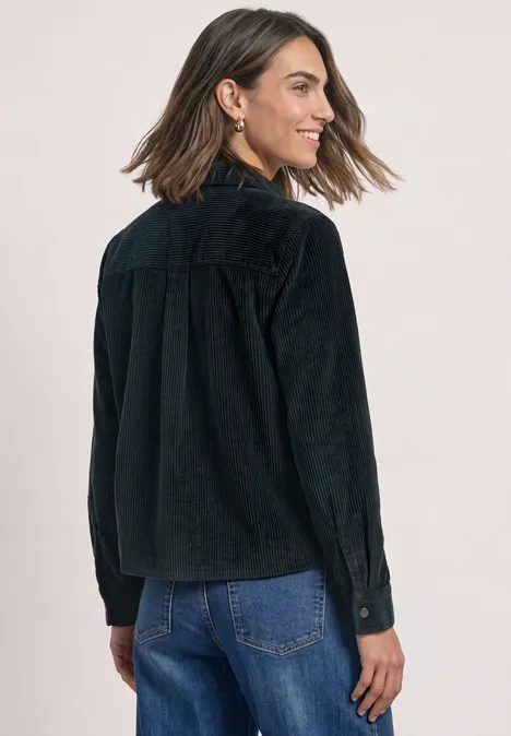 Frau mit schwarzer Kordjacke und blauen Jeans blickt lächelnd über die Schulter.