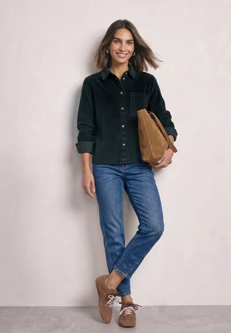 Frau in schwarzem Cordhemd, blauen Jeans, braunen Stiefeletten, trägt eine cognacfarbene Tasche.