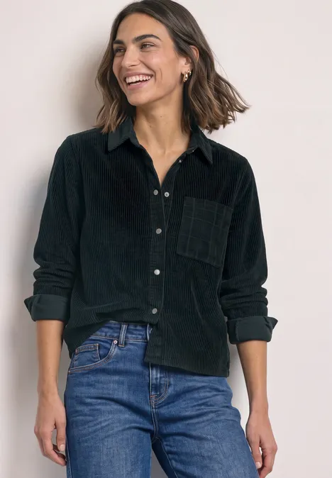 Frau in schwarzem Cord-Hemd und Jeans, lächelt vor einfachem, hellem Hintergrund.