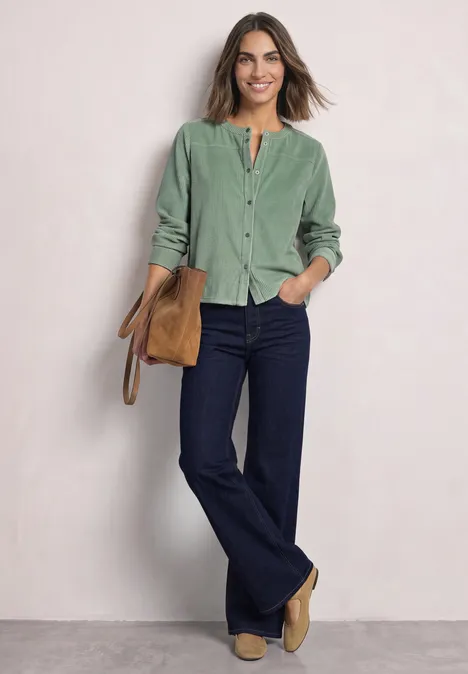 Eine Frau trägt ein grünes Cordhemd, dunkle Schlagjeans, beige Stiefeletten und hält eine Tasche vor hellem Hintergrund.