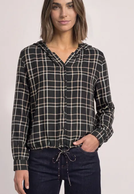 Frau in schwarzem kariertem Kapuzenhemd mit Tunnelzug am Saum und dunkler Jeans.