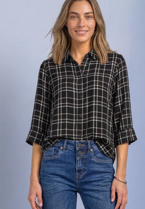 Femme souriante en chemise noire à carreaux et jean bleu sur fond neutre.