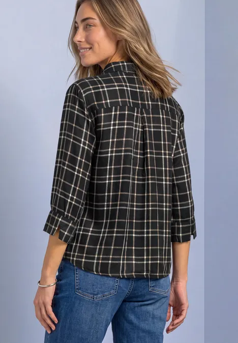 Femme de dos portant une chemise à carreaux noire à manches 3/4 et un jean bleu, souriante par-dessus l'épaule.