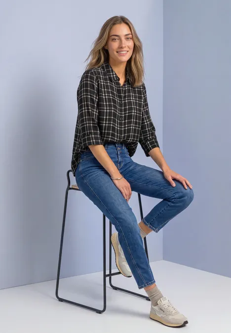 Femme souriante assise sur un tabouret haut, portant une chemise à carreaux, jean et baskets blanches.