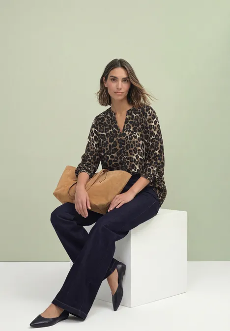 Eine Frau sitzt vor grünem Hintergrund auf weißem Block, trägt Leo-Bluse, dunkle Hose und hält eine beige Tasche.
