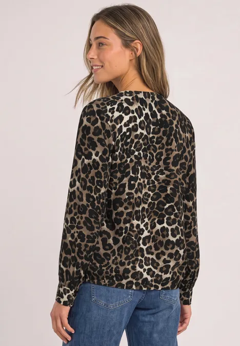 Junge Frau in seitlicher Pose trägt langärmlige Bluse im Leopardenmuster und blaue Jeans vor neutralem Hintergrund.