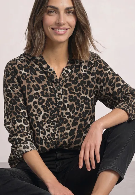 Eine lächelnde Frau mit braunen Haaren trägt eine Leoparden-Bluse und schwarze Hose vor hellem Hintergrund.