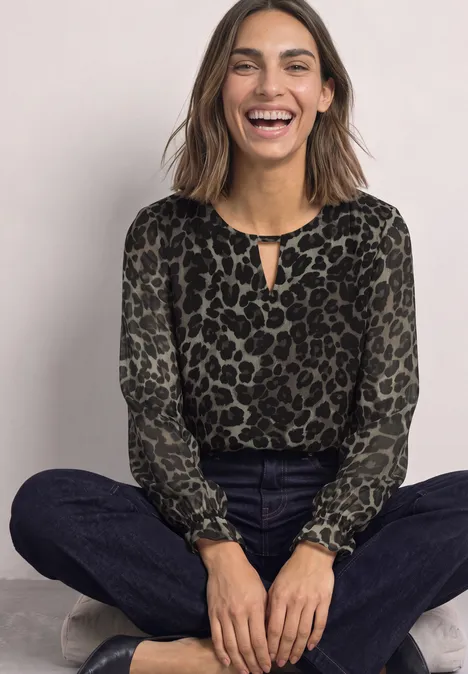 Lachende Frau sitzt crossbeinig in Leoparden-Print-Bluse, dunklen Jeans und Sneakers vor neutralem Hintergrund.