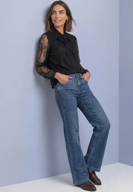 Langarm Bluse aus Spitze mit Schleifendetail Black Frau in schwarzer Spitzenbluse und weiten Jeans lehnt an heller Wand und trägt Stiefel.