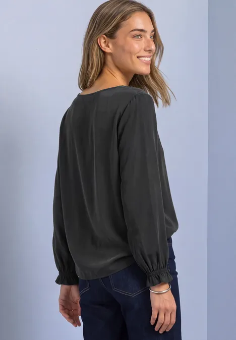 Blouse met ronde hals en gespdetail armor grey Vrouw kijkt over haar schouder, glimlacht en draagt een zwarte blouse met lange mouwen en een spijkerbroek.