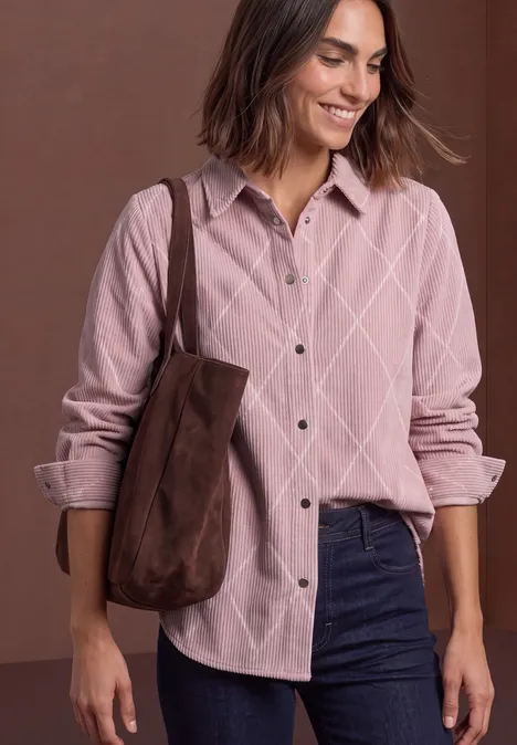 Frau in rosa Cordhemd mit Rautenmuster, dunkelblauen Jeans und brauner Schultertasche, lächelnd.