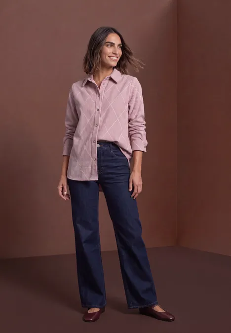 Frau in rosa Hemd, blauer Jeans und Loafers vor braunem Hintergrund lächelt entspannt.