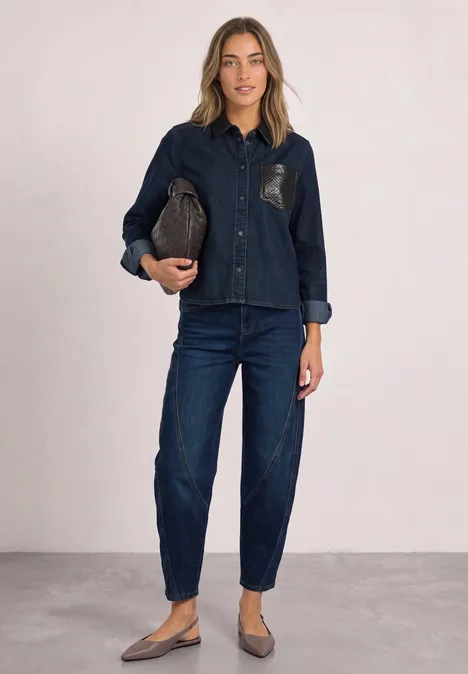 Eine Frau in dunklem Jeans-Hemd und -hose, hält eine braune Clutch und trägt flache Schuhe vor neutralem Hintergrund.