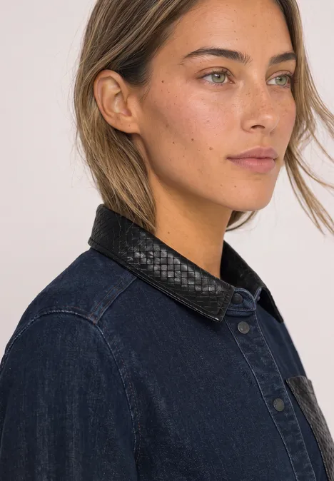 Eine Frau mit hellbraunem Haar in Jeansjacke mit schwarzem Lederkragen blickt seitlich vor neutralem Hintergrund.