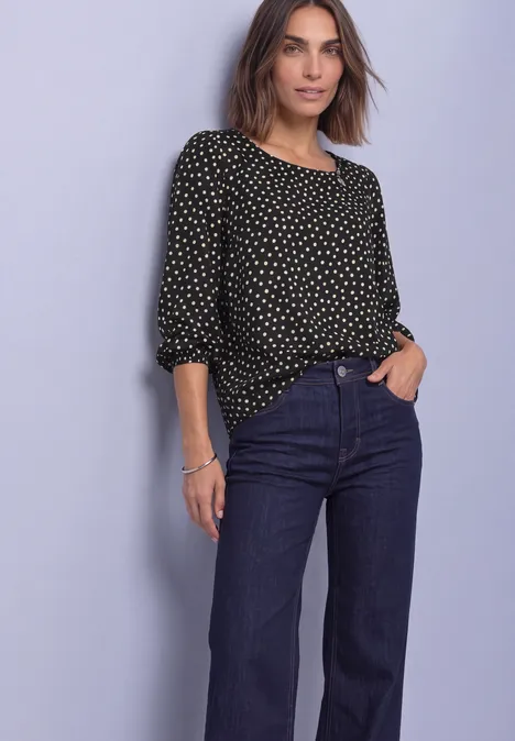 Vrouw in zwarte blouse met witte stippen en wijde donkerblauwe jeans tegen lichte achtergrond.