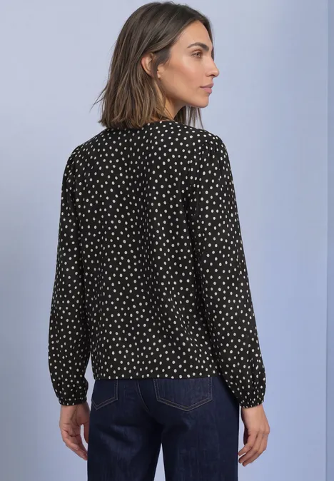 Vrouw van achteren in een zwart blouse met witte stippen en spijkerbroek, tegen een lichte achtergrond.