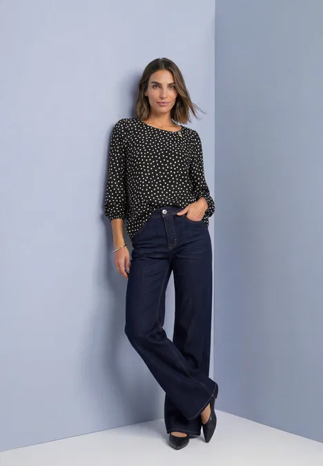 Vrouw in zwart polkadotblouse en wijdvallende jeans, nonchalant poserend voor een neutrale achtergrond.