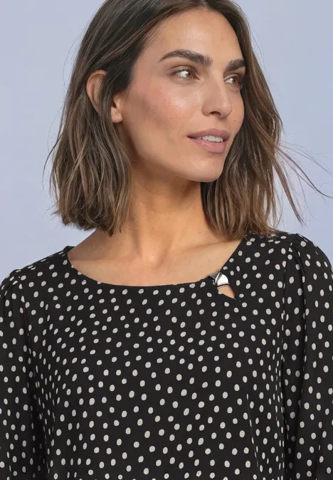Close-up van een vrouw met halflang bruin haar in een zwarte blouse met witte stippen.