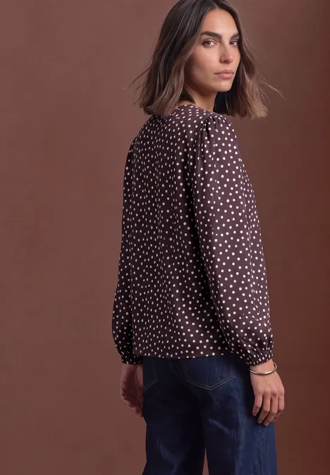 Een vrouw in een bruin blouse met witte stippen en een jeans, kijkt over haar schouder tegen een bruine achtergrond.