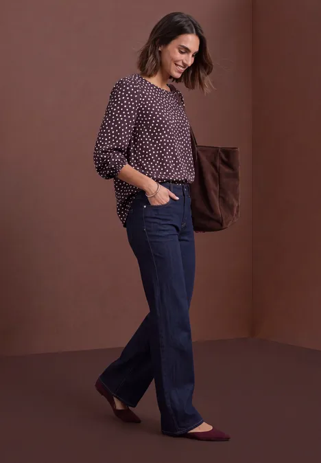 Vrouw lacht en loopt voor een bruine achtergrond, draagt wijdvallende spijkerbroek en een blouse met stippen.