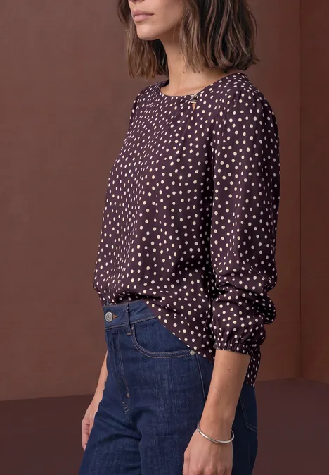 Een vrouw draagt een donkerblouse met witte stippen, een blauwe spijkerbroek en een armband voor een bruine achtergrond.