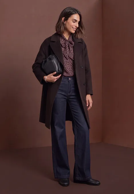 Femme en manteau marron, chemisier à motifs et jean large, tenant un sac noir.