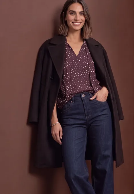 Femme souriante en manteau sombre, chemisier violet à pois et jean bleu, posant devant un fond marron.