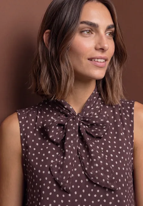 Femme en blouse marron sans manches à motif de coeurs blancs et nœud au col, fond brun.