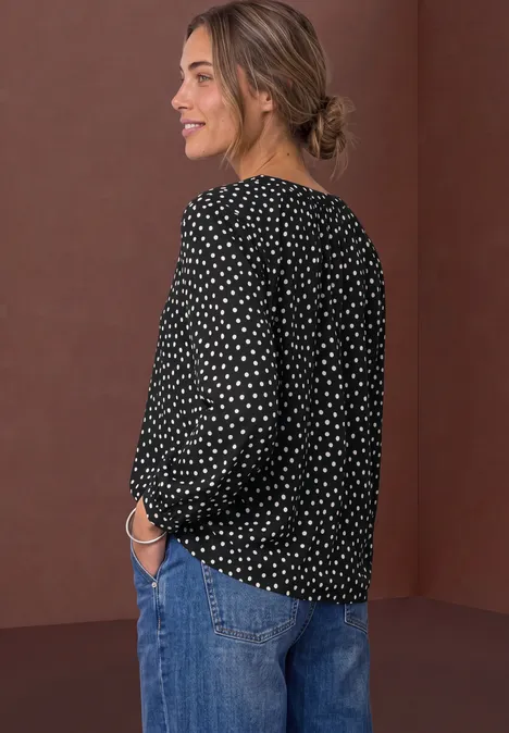 Frau im Profil trägt schwarze gepunktete Bluse und Jeans, lächelnd.