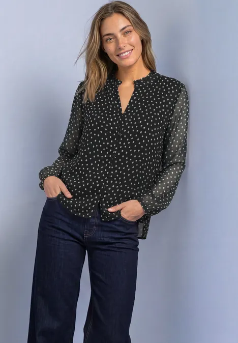 Een glimlachende vrouw draagt een zwarte bloes met stippen en een donkere jeans.