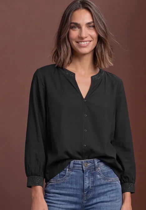 Een lachende vrouw in een zwart overhemd en jeans tegen een bruine achtergrond.