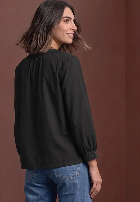 Rugaanzicht van een vrouw in een losse zwarte blouse met lange mouwen en jeans voor een bruine achtergrond.