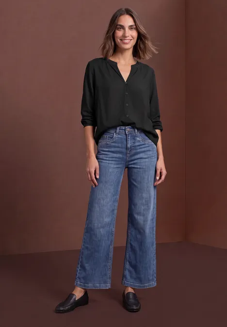 Vrouw in zwarte blouse en blauwe jeans voor een bruine achtergrond.