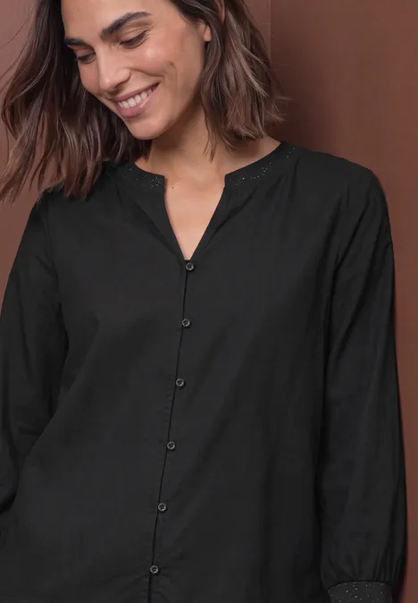 Een glimlachende vrouw in een zwarte blouse met knopen en lange mouwen, bruine achtergrond.