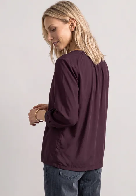 Blondynka ma na sobie fioletową bluzkę i dżinsy, stoi bokiem i patrzy w dół.