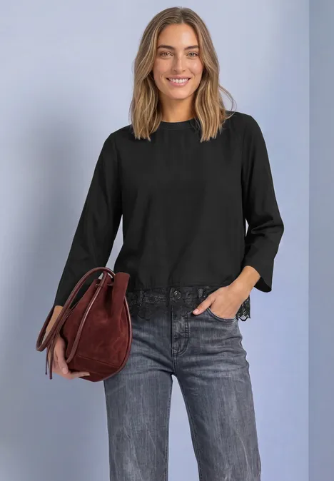 Frau in schwarzer Bluse mit Spitzenabschluss, grauen Jeans, hält roten Veloursleder-Beutel und lächelt.