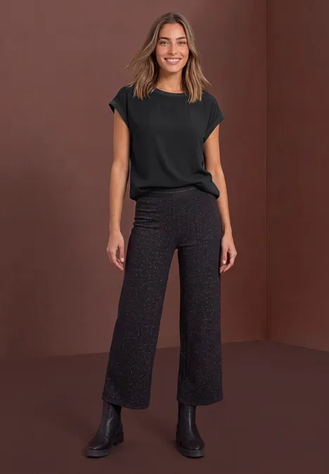Een vrouw in een zwart shirt en wijde, donkere broek voor een bruine achtergrond.
