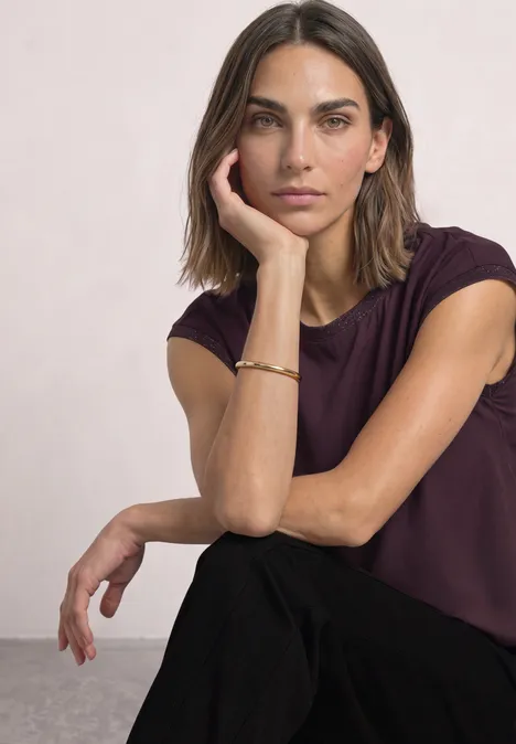 Une femme aux cheveux bruns porte un t-shirt violet, appuie sa tête sur sa main et regarde l'objectif.
