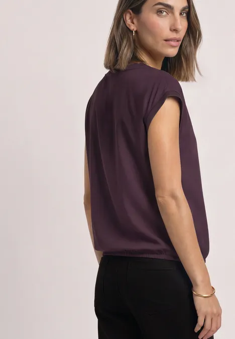 Une femme porte un top sans manches violet et un pantalon noir, regarde par-dessus l'épaule.