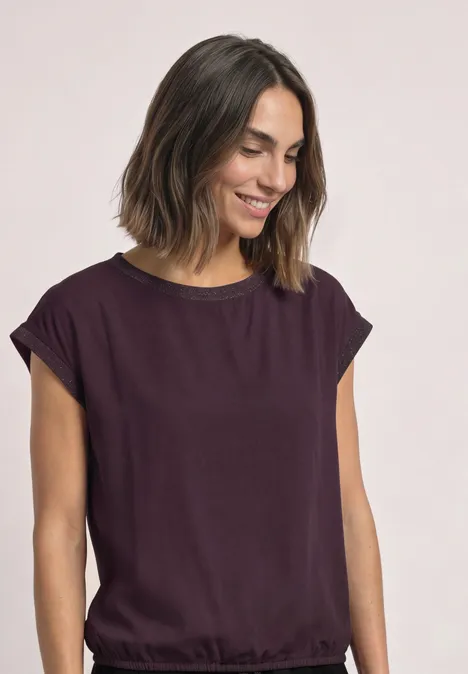 Femme dans un top violet ample, sourit en regardant vers le bas, fond neutre.