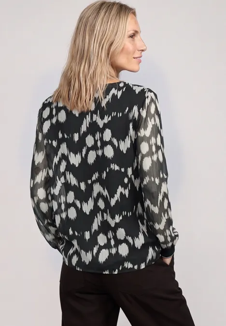 Chiffon Bluse mit Rundhals und Muster Black Frau in schwarzer Bluse mit weißem Muster, schaut seitlich. Schulterlange blonde Haare