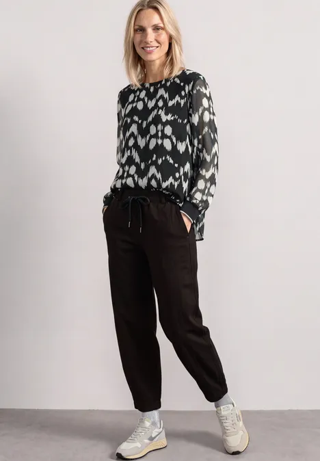 Chiffon Bluse mit Rundhals und Muster Black Frau trägt schwarze Jogginghose, gemustertes Longsleeve und weiße Sneaker.