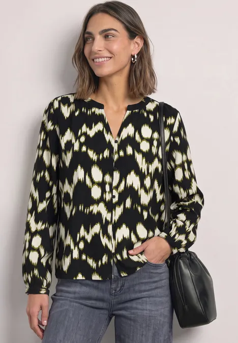 Vrouw met halflang haar draagt een zwart-geel-wit gemêleerde blouse, spijkerbroek en zwarte tas.
