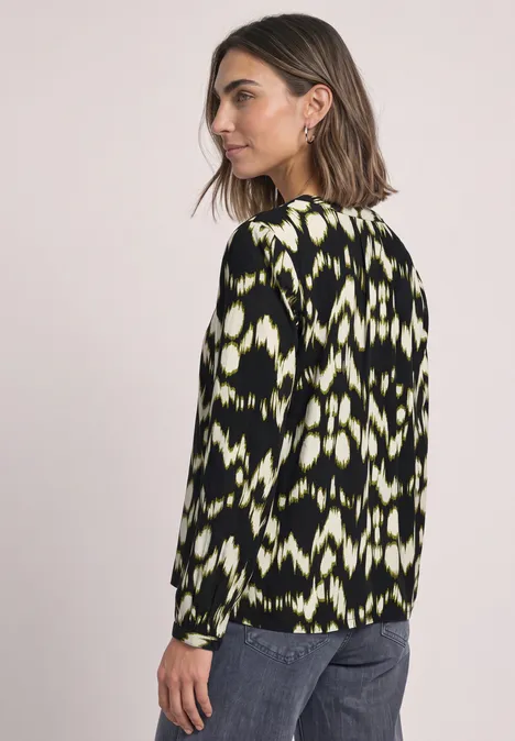 Vrouw in profiel met zwart langarmshirt in neongroen patroon en een spijkerbroek.