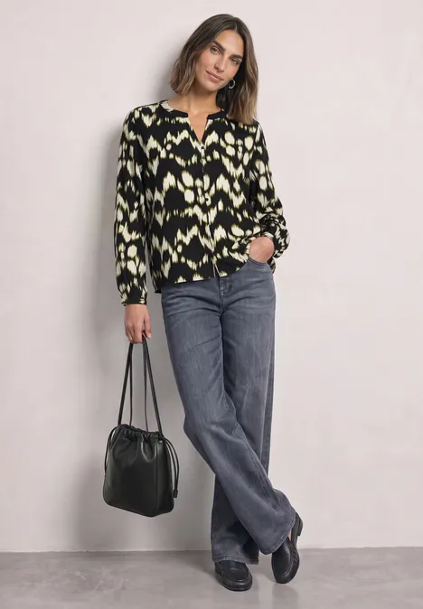 Een vrouw draagt een blouse met print, grijze jeans en zwarte schoenen en houdt een zwarte tas vast.